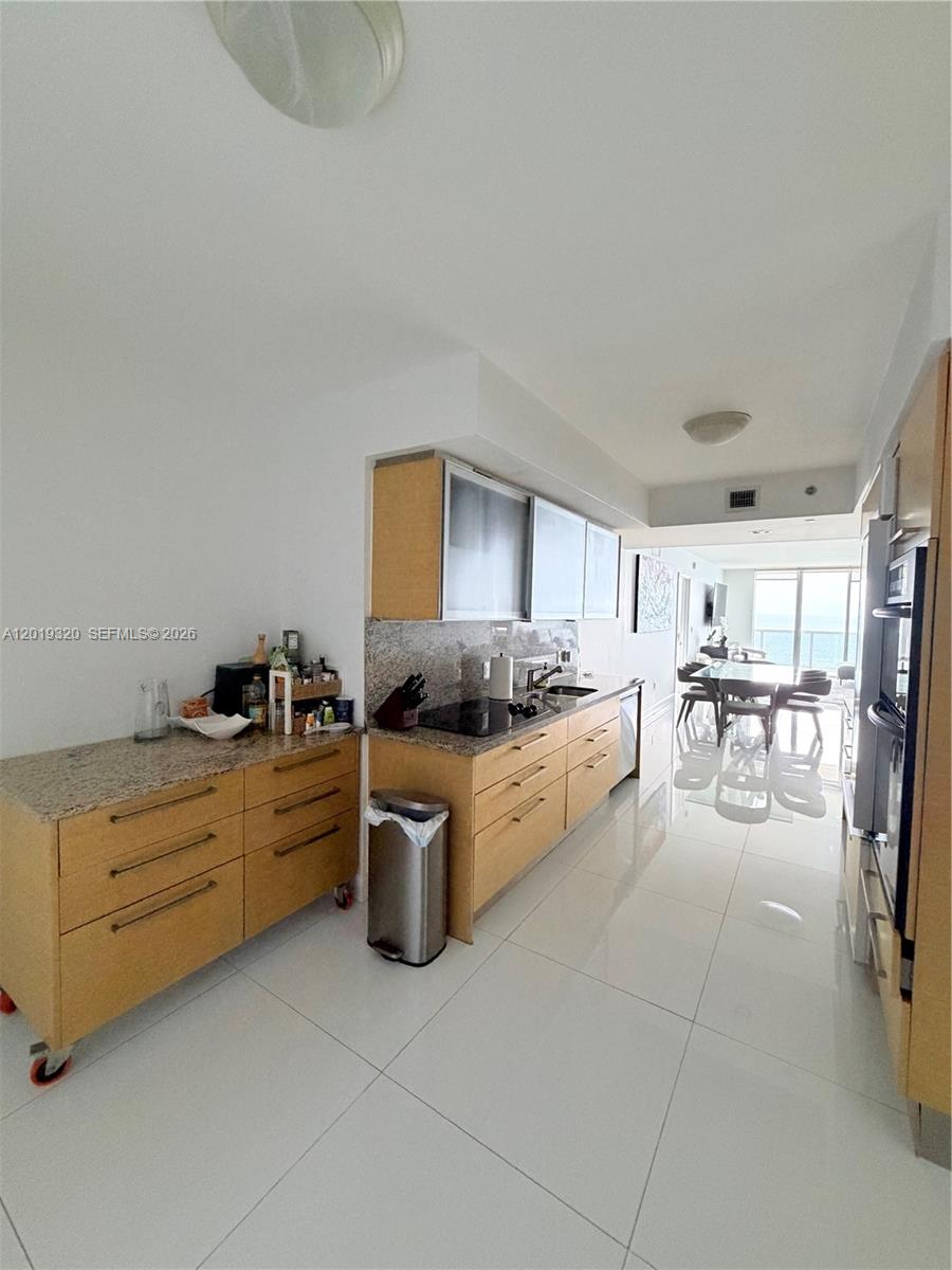 A modern kitchen features light wood cabinets, granite countertops, and white tile flooring — 1800 S Ocean Dr # 3002, Hallandale Beach FL 33009, HALLANDALE BEACH, FL