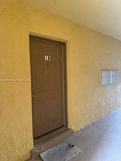A brown apartment door labeled 1B is set in a textured yellow exterior wall — 2832 NW 55th Ave # 1B, Lauderhill FL 33313, LAUDERHILL, FL