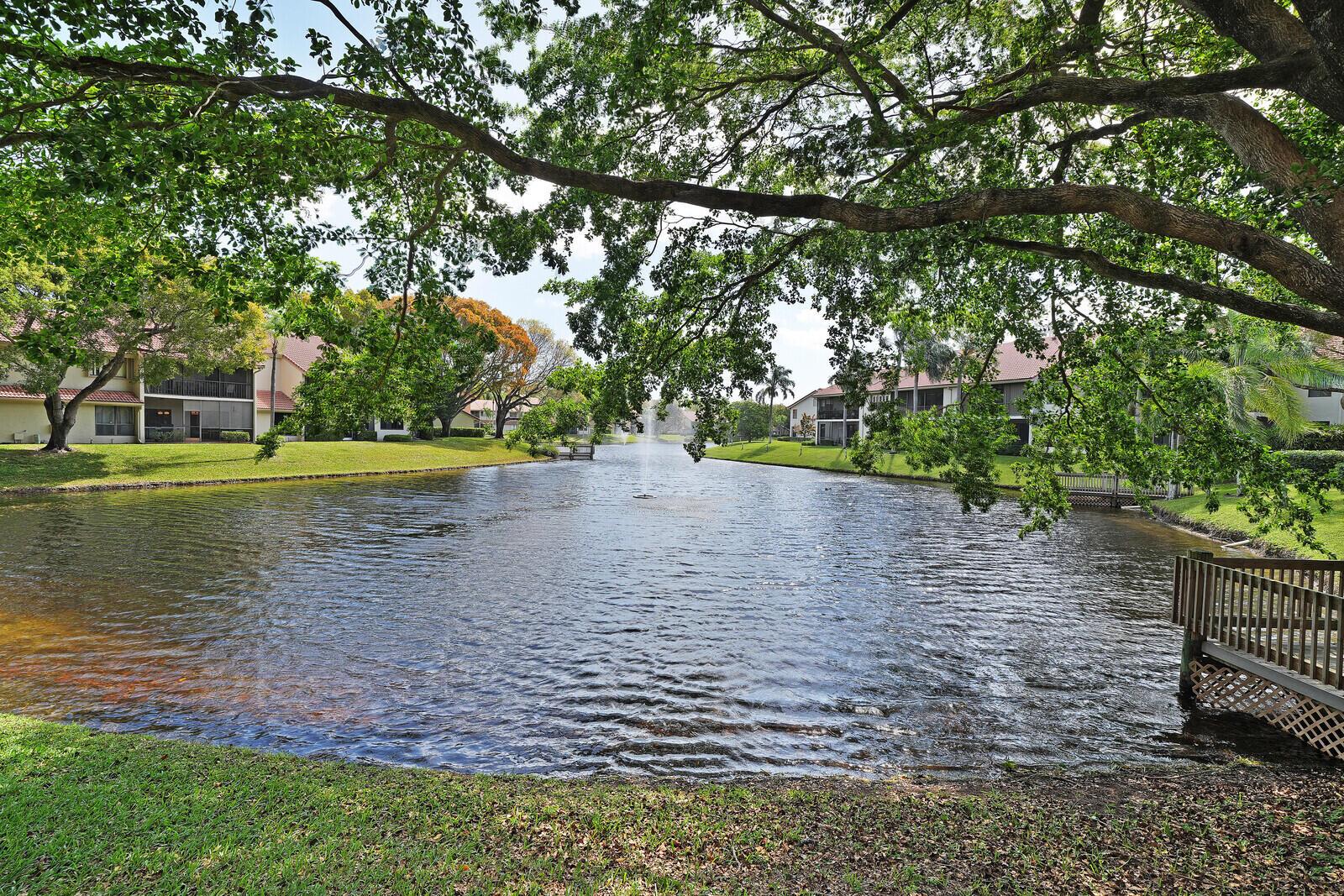 A view of a lake surrounded by apartment buildings, green lawns, and large shade trees — 5521 Coach House Circle # D, Boca Raton FL 33486, BOCA RATON, FL