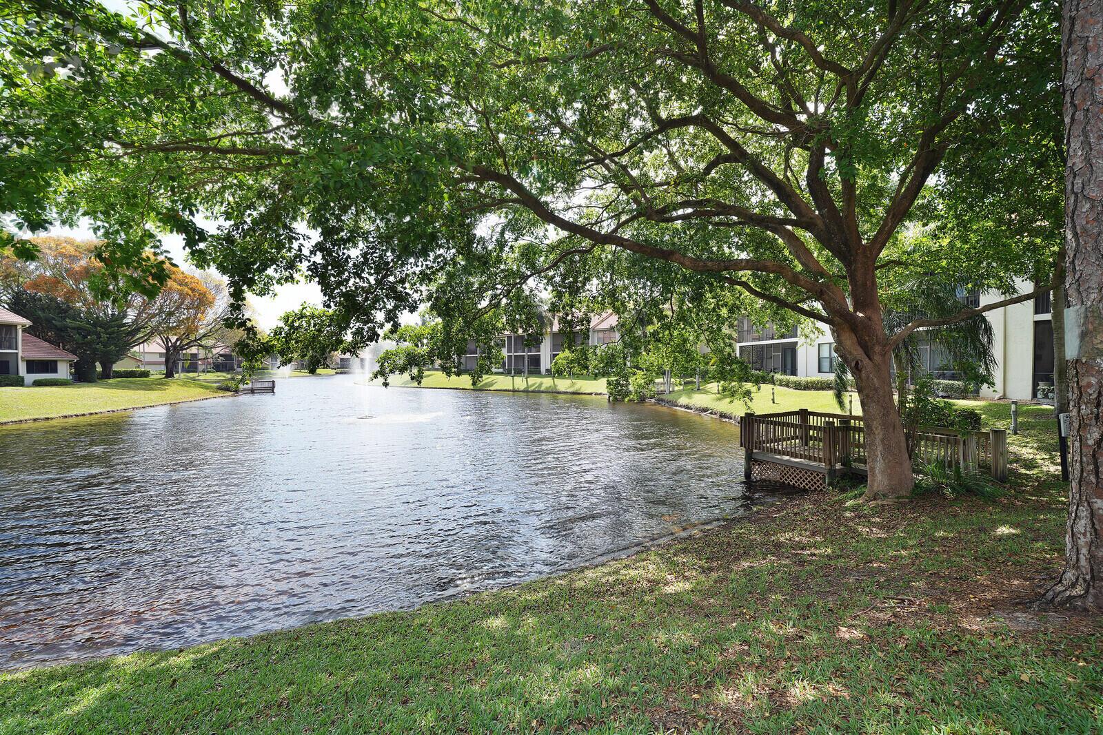 A small wooden deck sits on the grassy bank of a pond with a fountain — 5521 Coach House Circle # D, Boca Raton FL 33486, BOCA RATON, FL