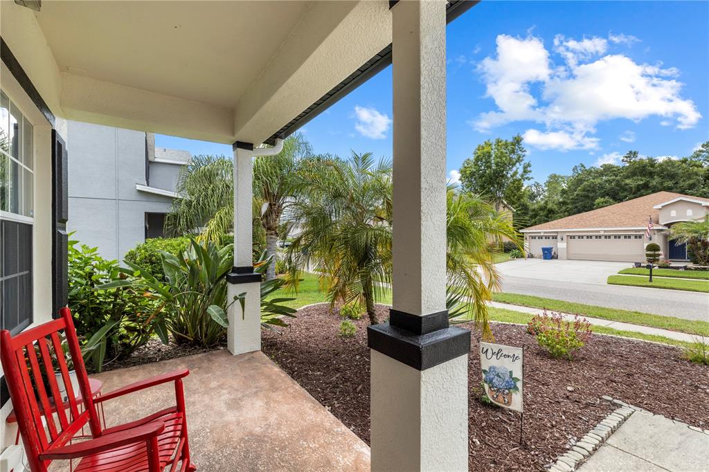A covered front porch features a red rocking chair and a welcome sign in the yard — 6053 EVERLASTING PLACE, LAND O LAKES, FL