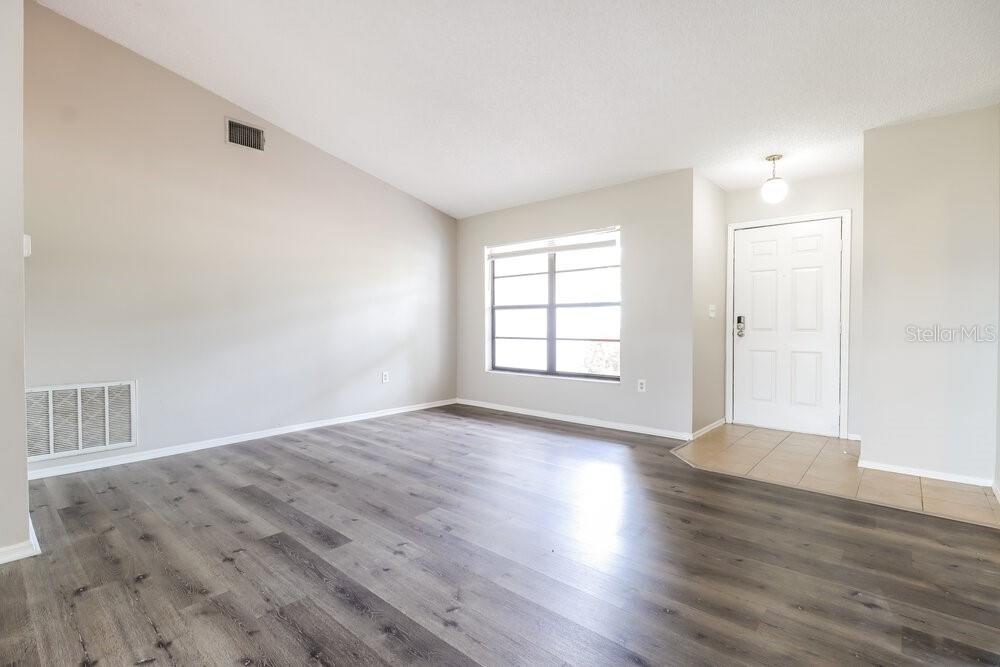 An empty living room features gray wood-look flooring, a vaulted ceiling, and a front door — 2225 HIDDEN MEADOWS DRIVE E, PALM HARBOR, FL