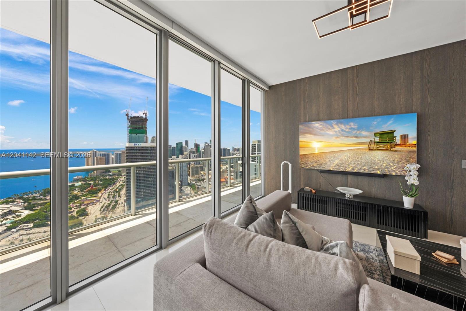 A modern living room features floor-to-ceiling windows overlooking a city skyline and ocean — 888 Biscayne Blvd # 5404, Miami FL 33132, MIAMI, FL