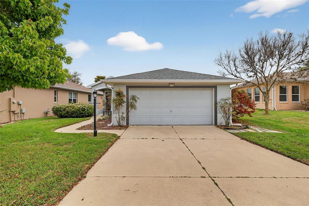 A single-story house with a two-car garage and a concrete driveway under a cloudy sky — 27106 GREENFLY ORCHID LANE, LEESBURG, FL