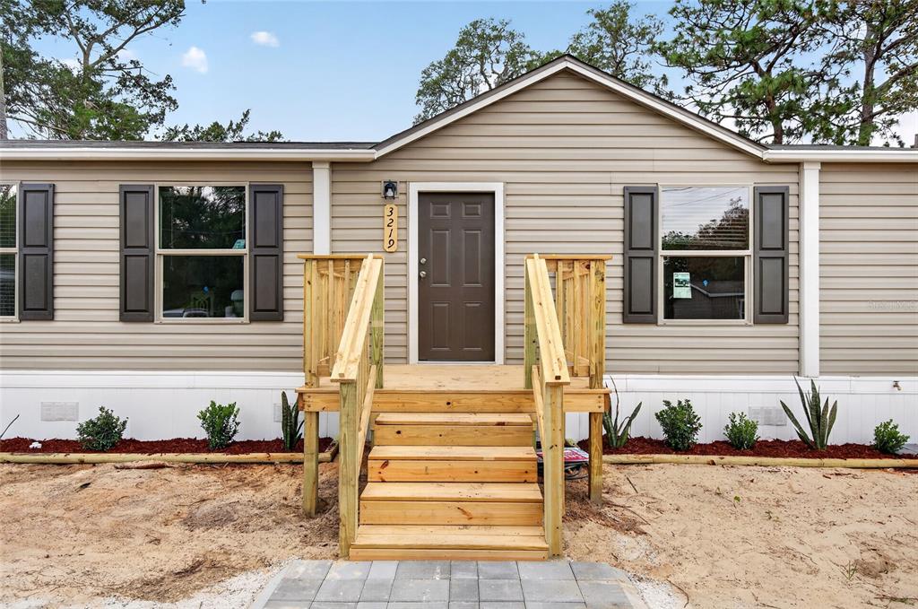 A tan mobile home features a wooden front porch with stairs and dark brown shutters — 3219 WELSH STREET, SPRING HILL, FL