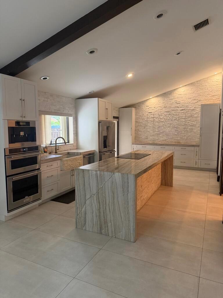 A modern kitchen features a large stone waterfall island, white cabinetry, and textured stone walls — 9553 Majestic Way, Boynton Beach FL 33437, BOYNTON BEACH, FL