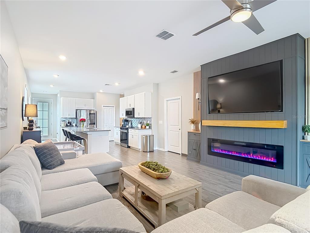 A living room with a gray fireplace wall and TV opens into a white kitchen — 7531 ASHCROFT DRIVE, WESLEY CHAPEL, FL