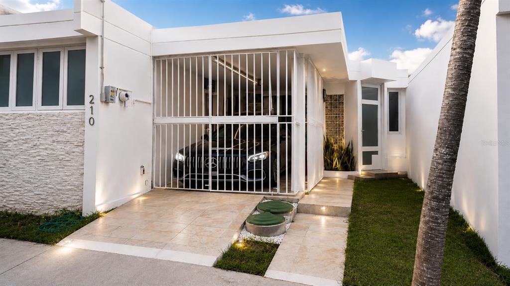 A modern white house exterior features a gated carport, a glass-paneled door, and landscaping — 210 CALLE FIRMAMENTO, CAROLINA, PR