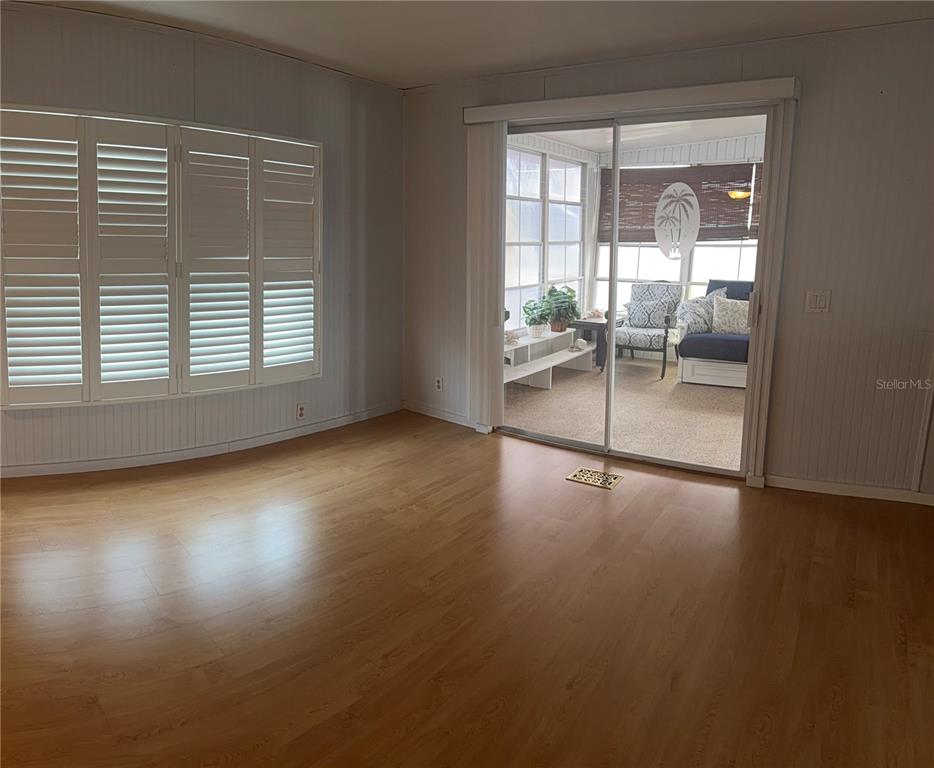A living room with light wood flooring, plantation shutters, and a sliding door to a sunroom — 3301 ALT 19 UNIT 130, DUNEDIN, FL