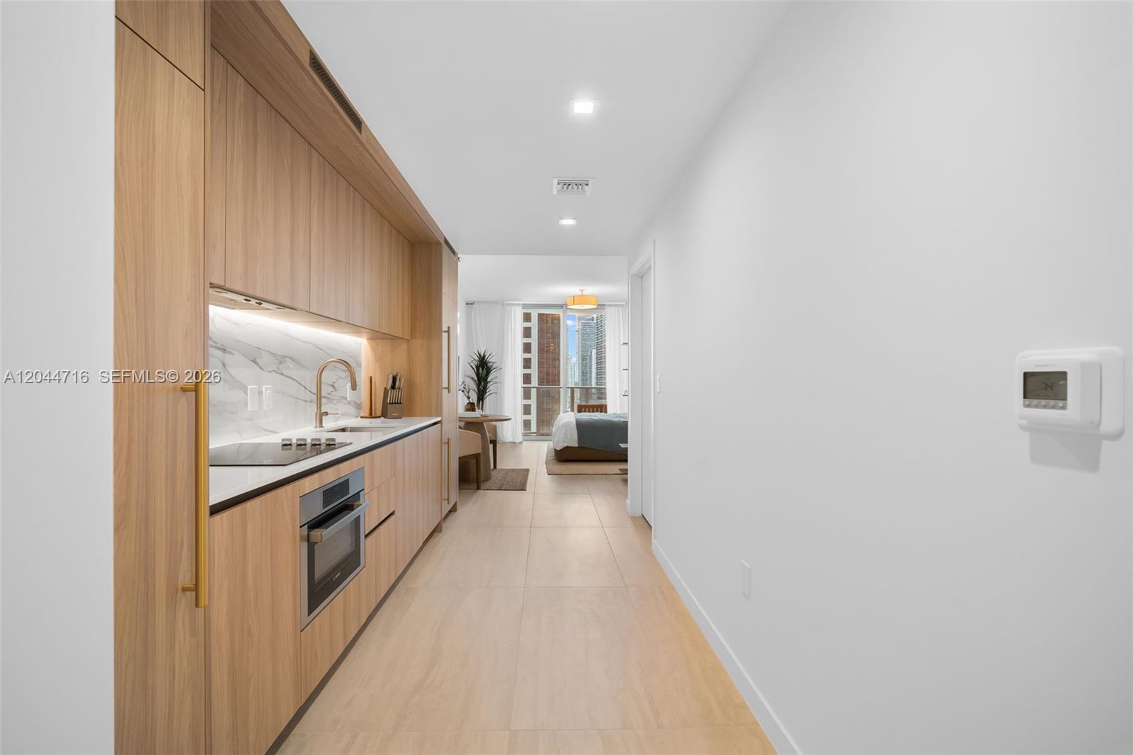 A modern kitchen with light wood cabinetry leads into a bedroom with a city view — 77 SE 5 Street # 2313, Miami FL 33131, MIAMI, FL