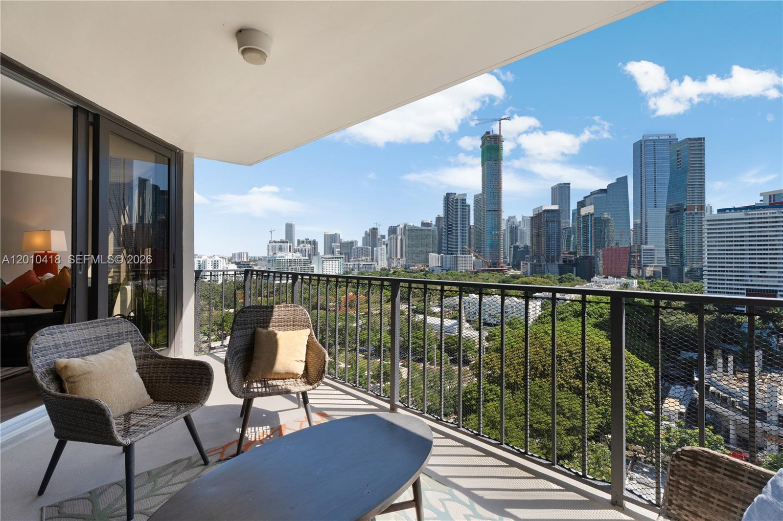 A balcony with wicker furniture overlooks a city skyline, construction sites, and the ocean — 1901 Brickell Ave # B1809, Miami FL 33129, MIAMI, FL