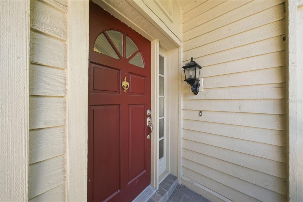 A red front door with a glass fanlight sits beside a wall-mounted outdoor light fixture — 50 PELICAN PLACE UNIT 50, CLEARWATER, FL