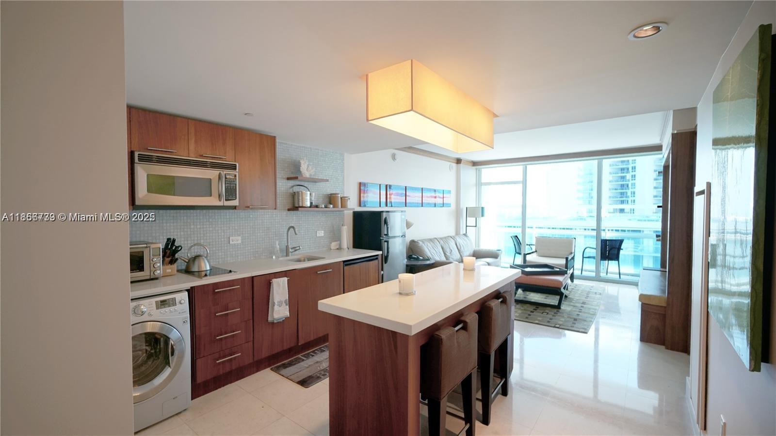 A modern kitchen with wood cabinets and a white island opens into a living area — 6801 Collins Ave # 710, Miami Beach FL 33141, MIAMI BEACH, FL