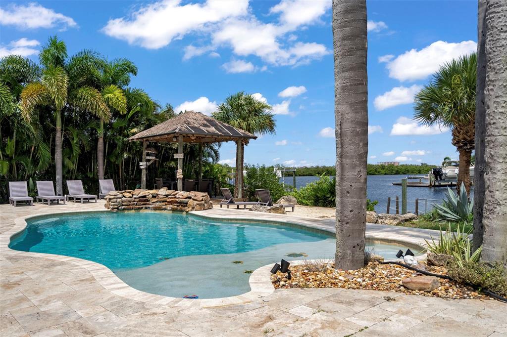 A swimming pool with a rock waterfall and a thatched-roof gazebo overlooks a waterfront view — 5548 LEEWARD LANE, NEW PORT RICHEY, FL