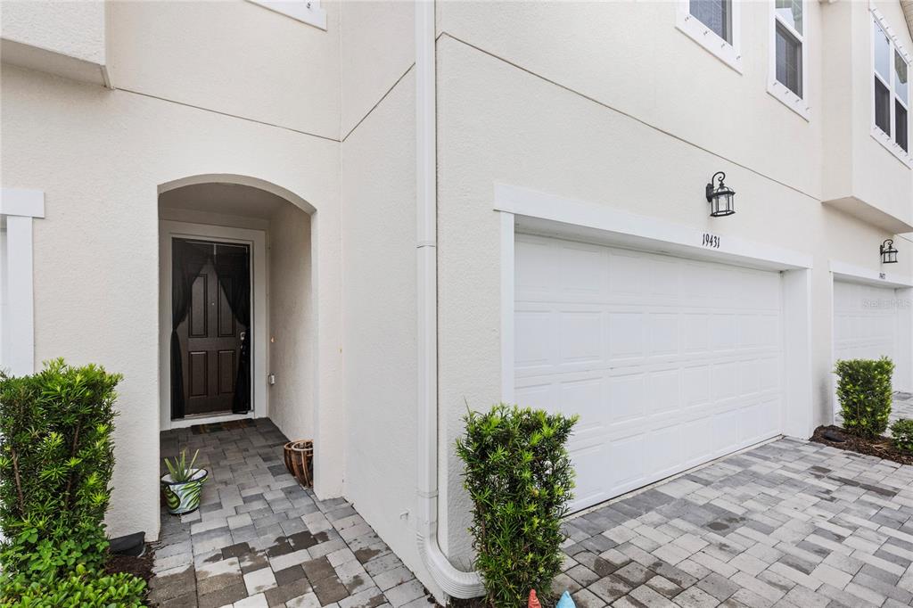A beige townhouse exterior features a white garage door, a covered entryway, and paver driveway — 19431 MONARCH WIND WAY, LUTZ, FL
