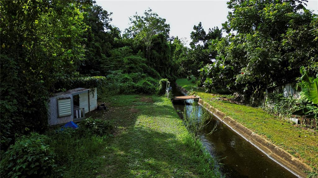 A small shed sits on a grassy lawn next to a narrow concrete drainage canal — Calle Brillante ARSENALES BAJOS, ISABELA, PR