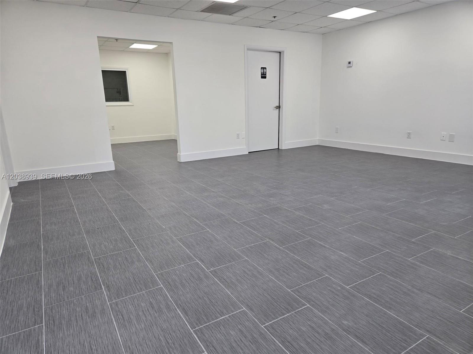 An empty office space features gray tile flooring, white walls, and a restroom door — HIALEAH, FL
