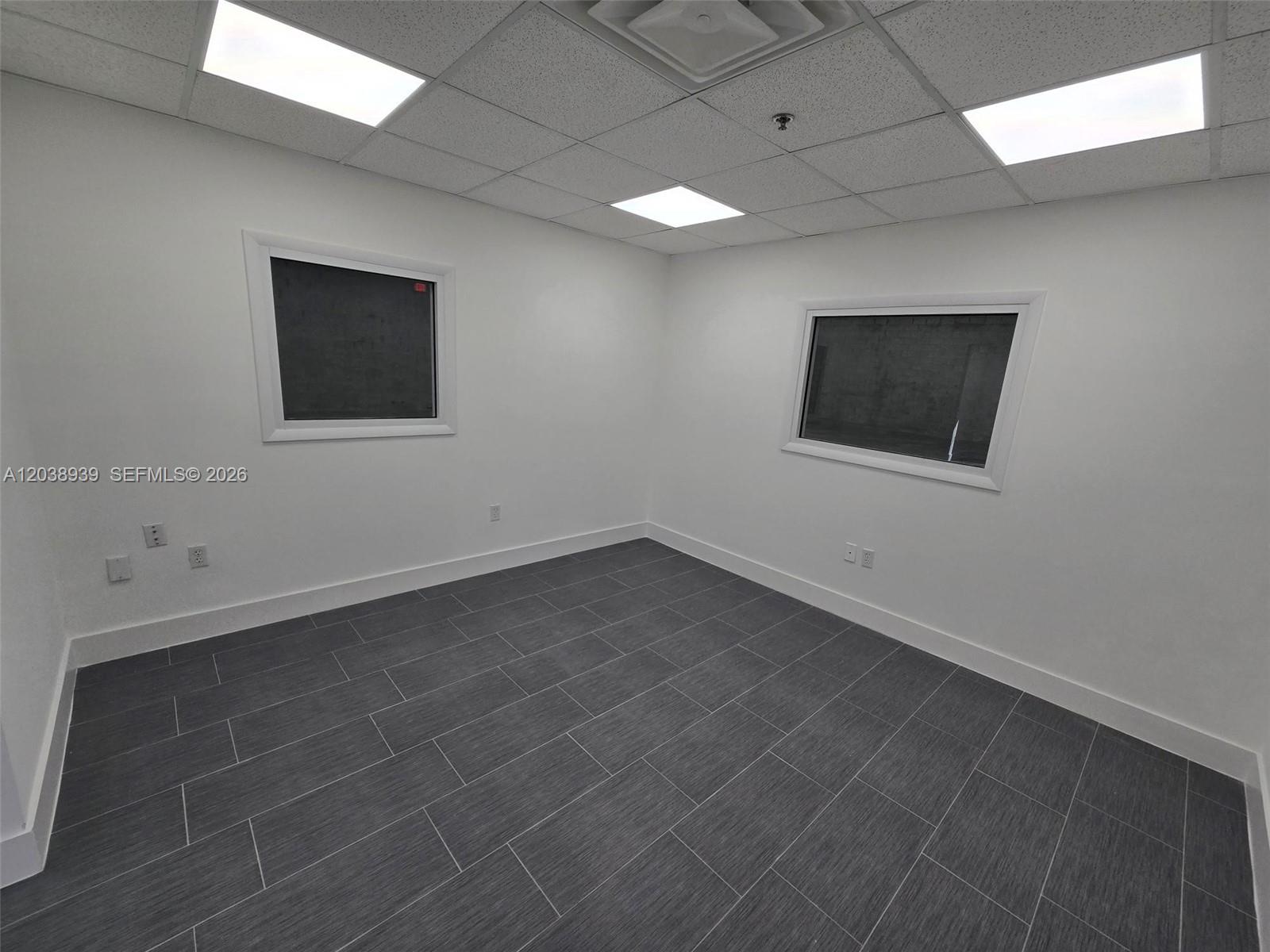 An empty office room features white walls, dark gray tile flooring, and two windows — HIALEAH, FL