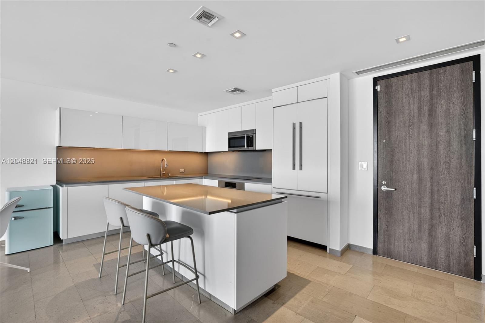 A modern kitchen features white cabinetry, a central island with bar stools, and stone flooring — 68 SE 6th St # 3710, Miami FL 33131, MIAMI, FL