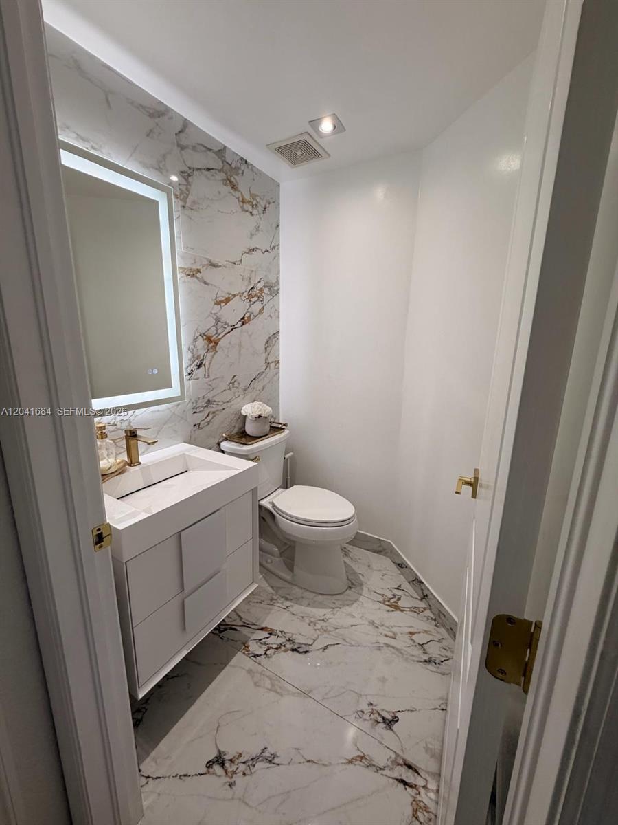 A small bathroom features a white vanity, a toilet, and marble-patterned tile flooring and walls — 10958 NW 86th Ter, Doral FL 33178, DORAL, FL