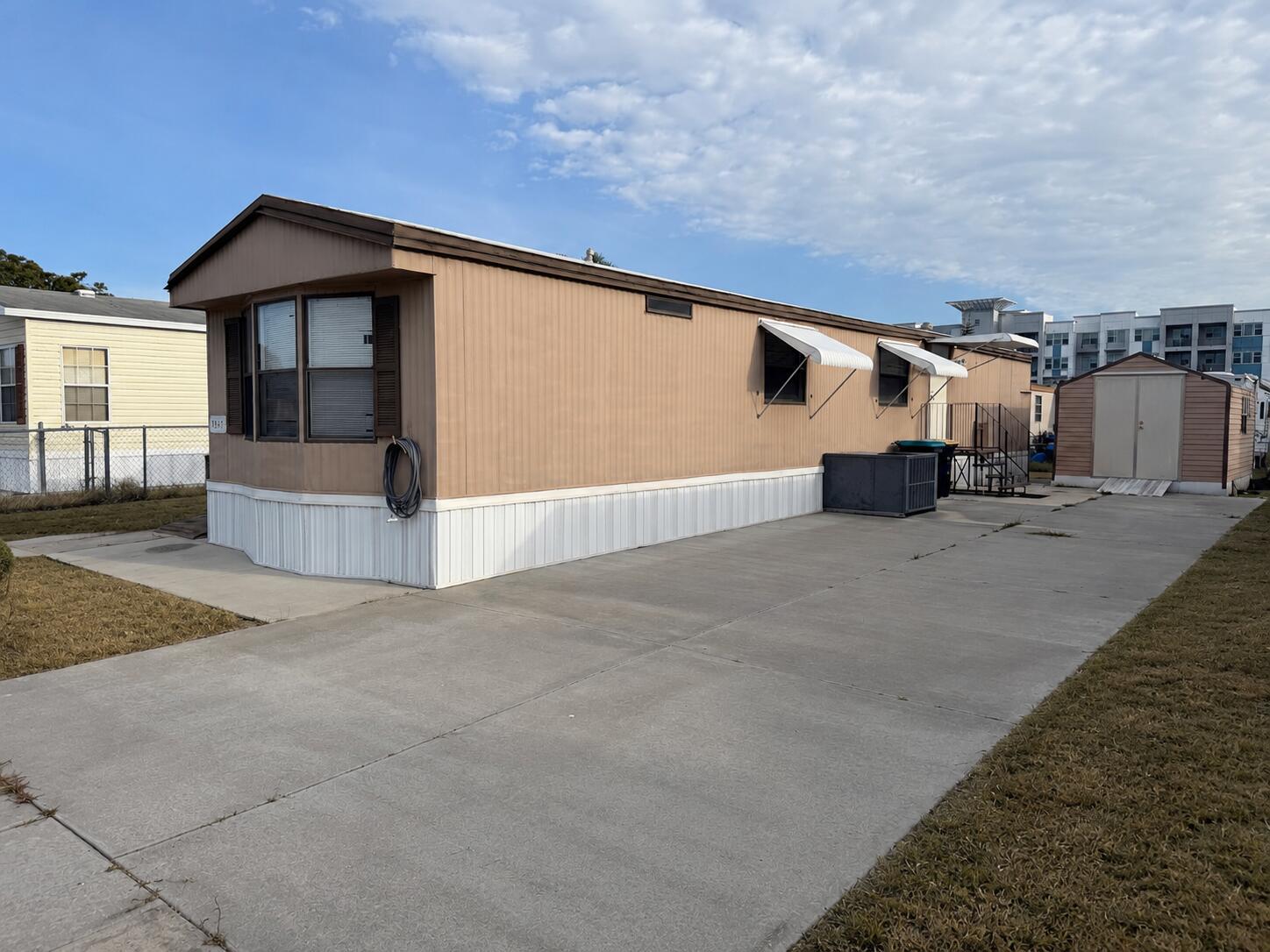 A tan mobile home with white skirting and a concrete driveway sits next to a shed — 2547 Chatham Way NE, Palm Bay FL 32905, PALM BAY, FL