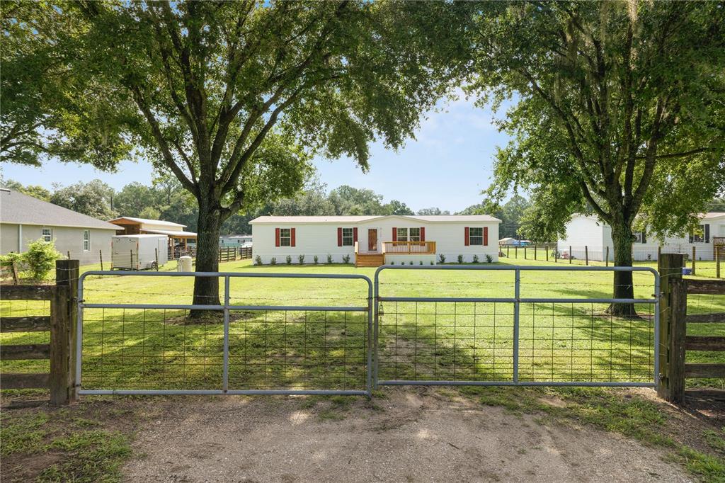 A single-story house sits behind a metal farm gate and large trees on a grassy lot — 7103 GLEN MEADOW DRIVE, LAKELAND, FL