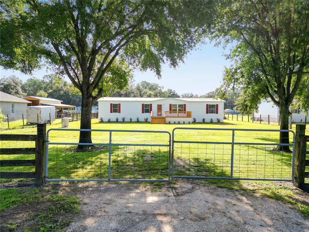 A white manufactured home with red shutters sits behind a metal gate and large trees — 7103 GLEN MEADOW DRIVE, LAKELAND, FL