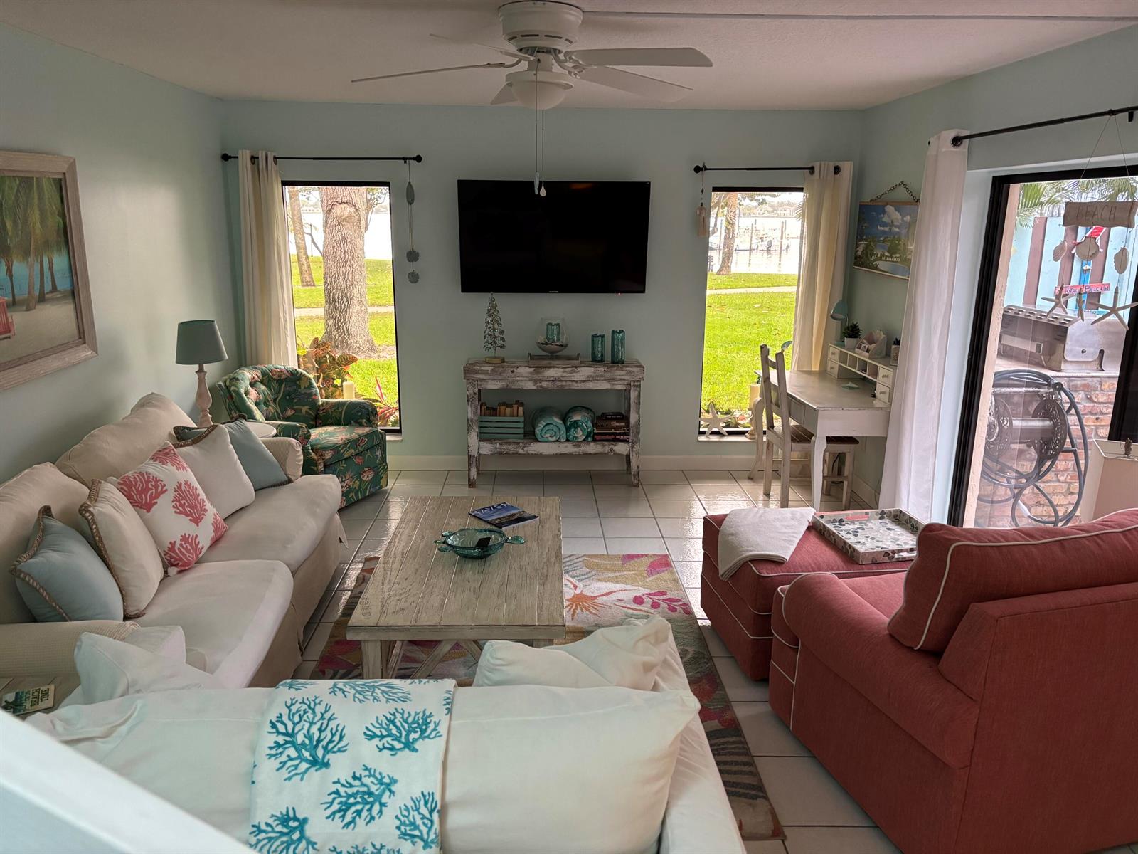 A living room features a white sofa, coral armchair, television, and large windows overlooking greenery — 825 Center Street # 21b, Jupiter FL 33458, JUPITER, FL