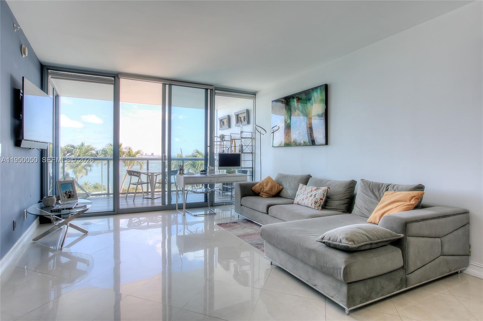 A living room features a grey sectional sofa, tile floors, and a balcony with water views — 601 NE 36th St # 710, Miami FL 33137, MIAMI, FL