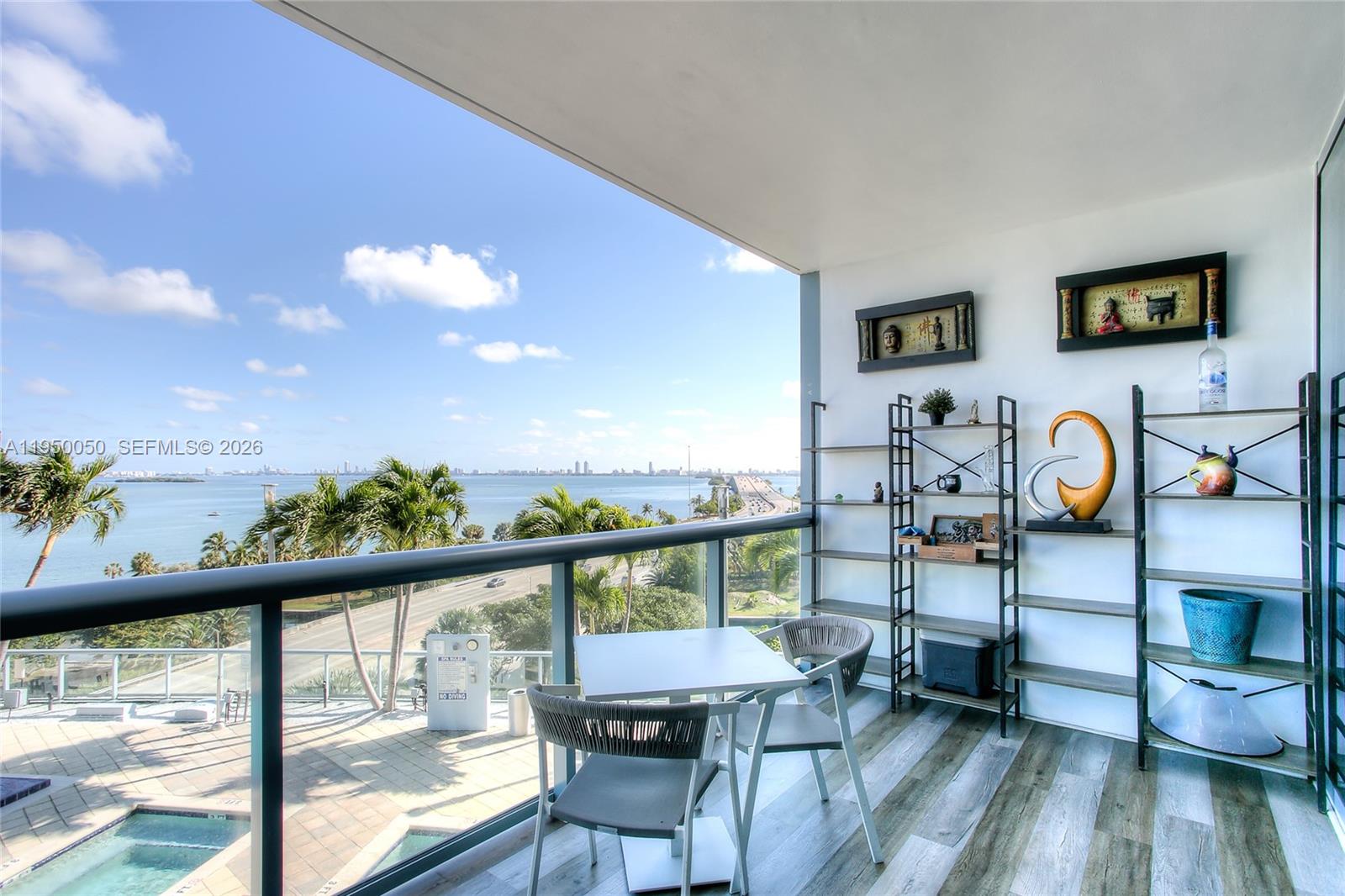 A balcony with a table, chairs, shelving, and a view of the water and bridge — 601 NE 36th St # 710, Miami FL 33137, MIAMI, FL