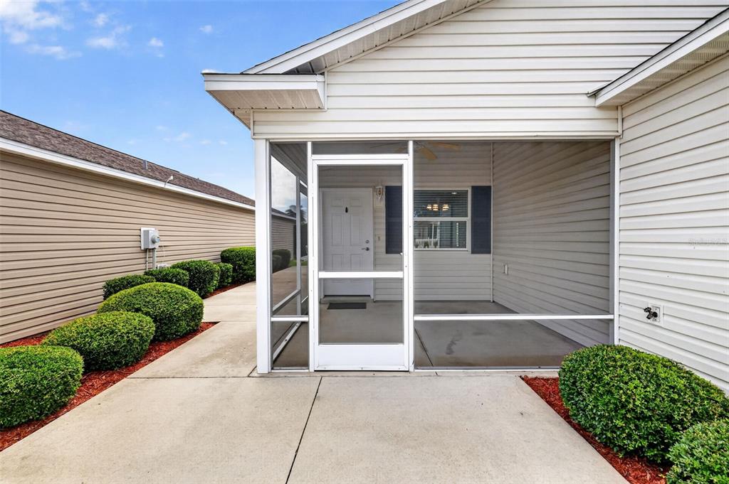 A screened-in porch with a white door and concrete patio sits beside a landscaped walkway — 3419 CARRABELLE COURT, THE VILLAGES, FL