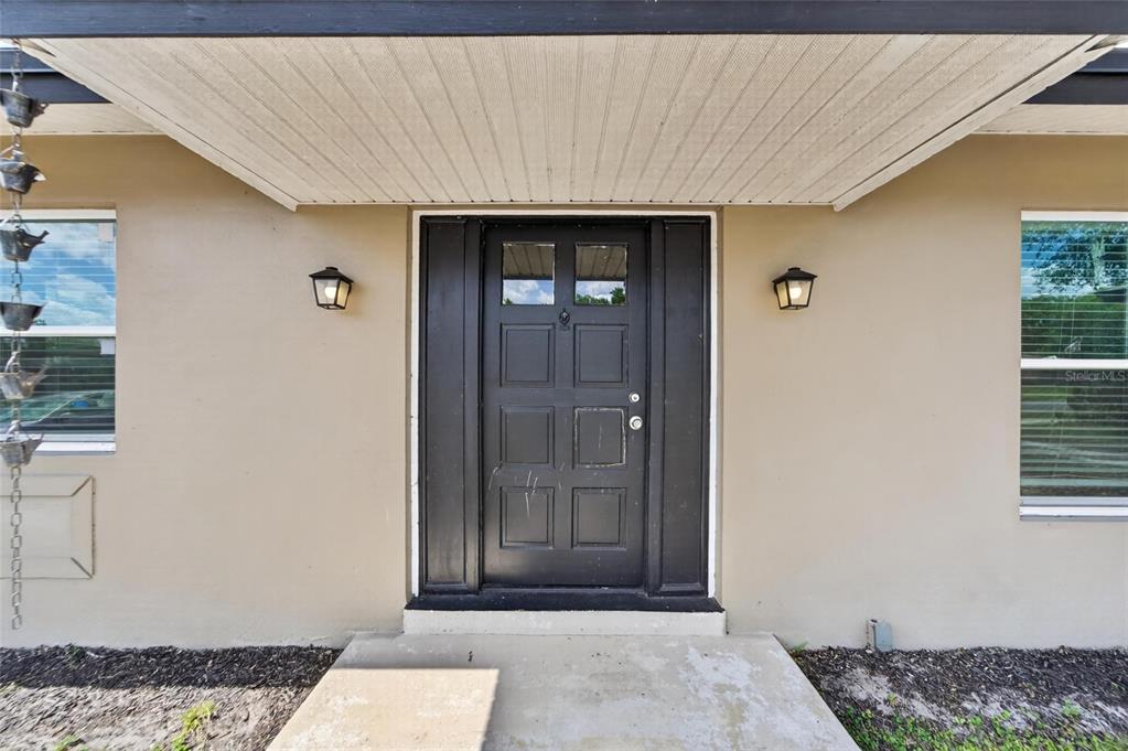A black front door with two windows is centered between two windows on a beige wall — 7156 LOCKWOOD STREET, SPRING HILL, FL