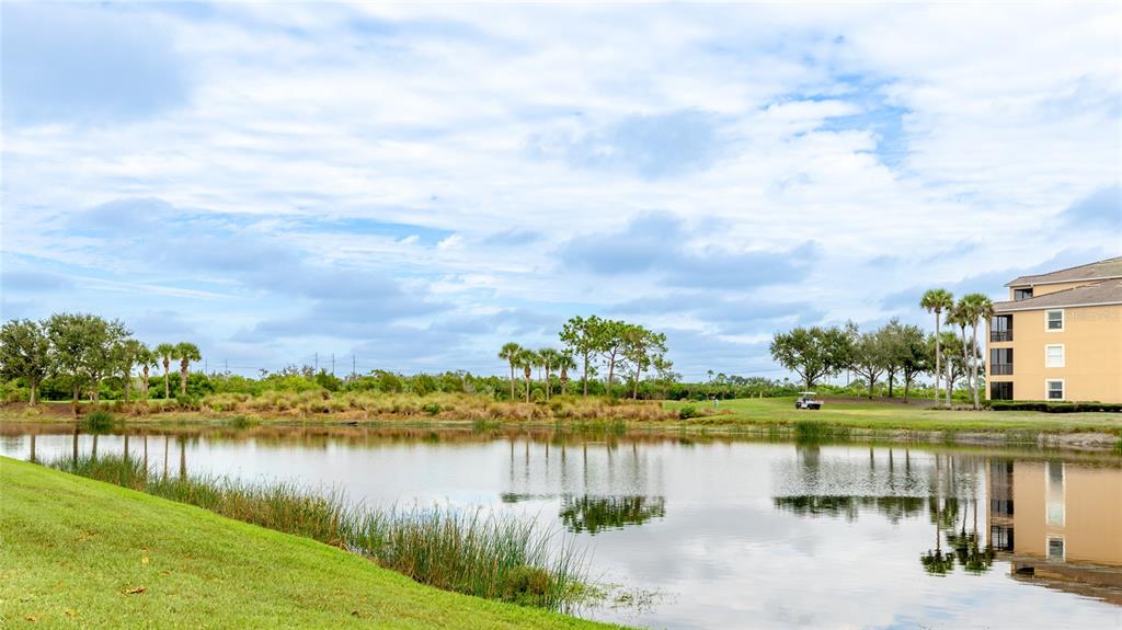 A pond sits in front of a multi-story building with palm trees and a golf cart — 7911 GRAND ESTUARY TRAIL UNIT 104, BRADENTON, FL