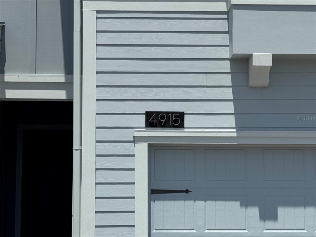 A light blue house exterior features a black address plaque with the number 4915 — 4915 TRIBUTE TRAIL, KISSIMMEE, FL
