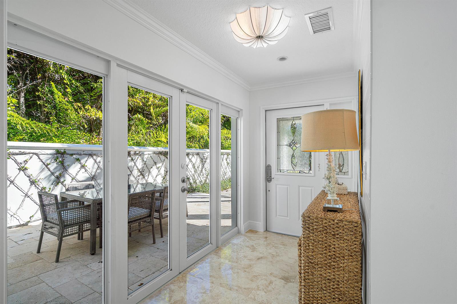 A bright entryway features a woven console table, a decorative lamp, and glass doors opening outdoors — 820 Palm Trail, Delray Beach FL 33483, DELRAY BEACH, FL