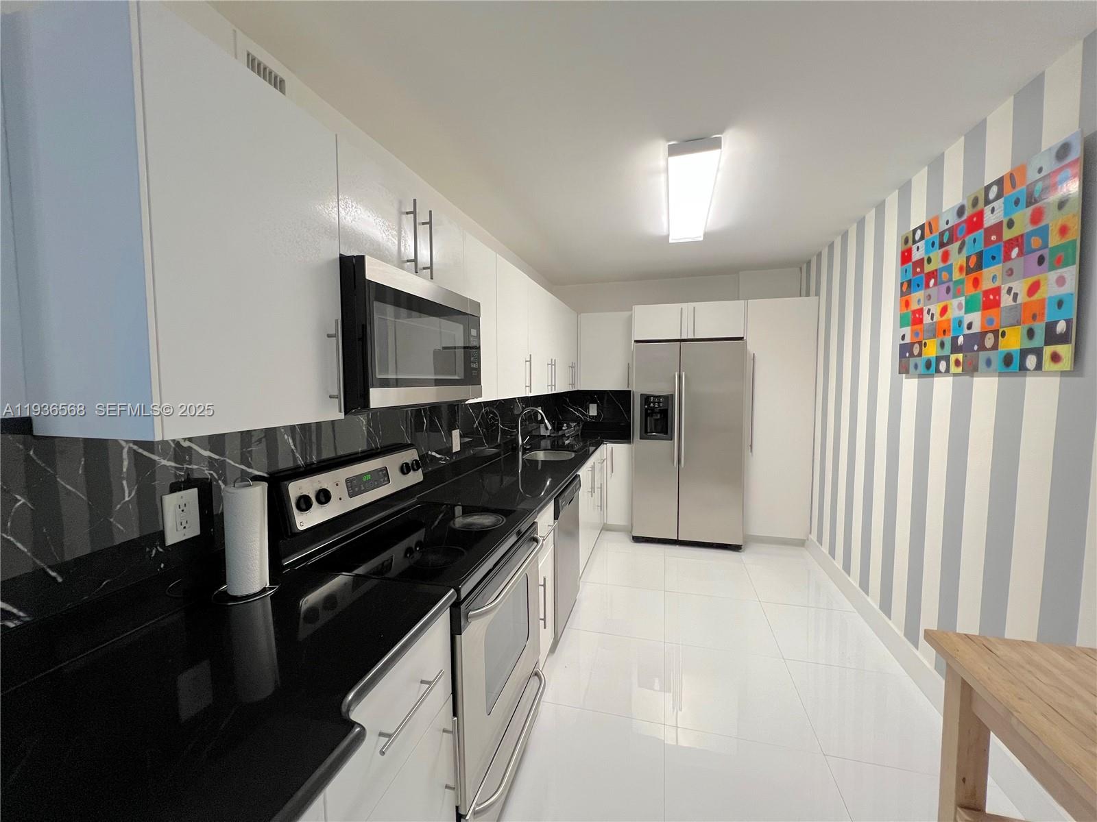 A galley kitchen features white cabinets, black countertops, a marble backsplash, and striped wallpaper — 401 69th St # 304, Miami Beach FL 33141, MIAMI BEACH, FL