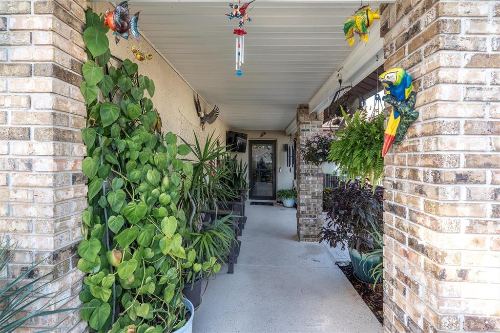 A covered brick porch features hanging plants, decorative animal figures, and a potted climbing vine — 7099 POND VIEW COURT, SPRING HILL, FL