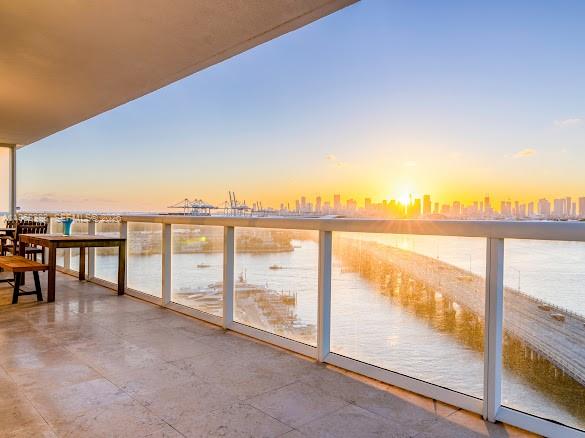 A balcony with a stone floor and glass railing overlooks a city skyline at sunset — 450 Alton Rd # 1202, Miami Beach FL 33139, MIAMI BEACH, FL