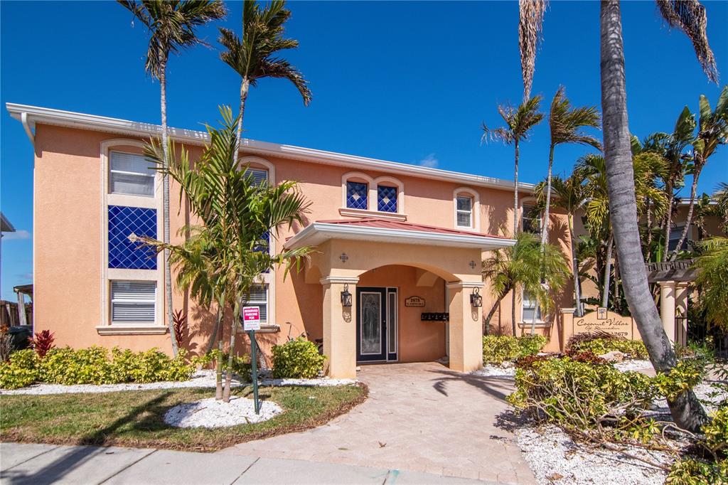 A two-story peach-colored apartment building features a covered entryway and palm trees in front — 2675 ST JOSEPHS DRIVE E UNIT B, DUNEDIN, FL