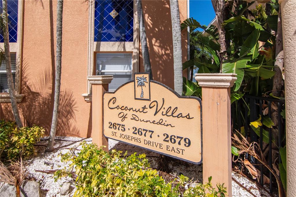A sign for Coconut Villas stands in front of a stucco building with blue windows — 2675 ST JOSEPHS DRIVE E UNIT B, DUNEDIN, FL