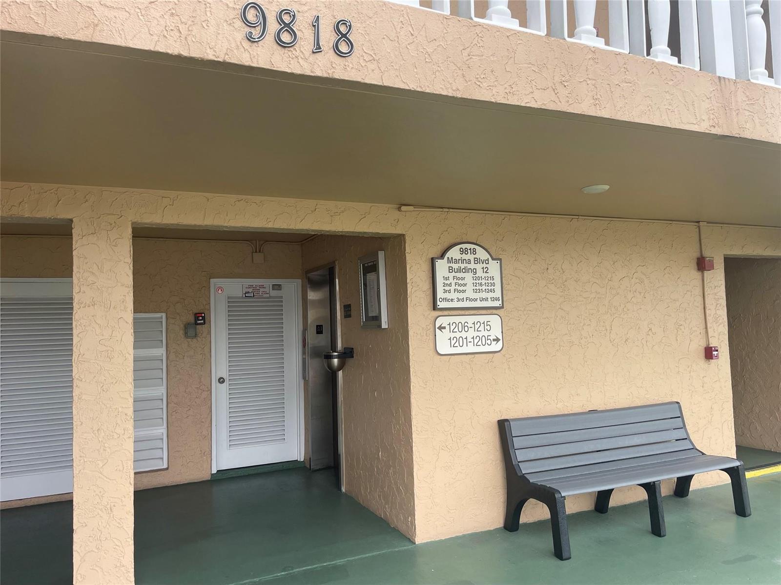 An exterior view of a building entrance with a bench, signage, and a white door — 9818 Marina Boulevard # 1220, Boca Raton FL 33428, BOCA RATON, FL