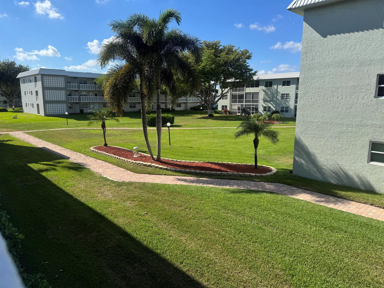 A grassy courtyard with palm trees and brick walkways between two multi-story apartment buildings — 9818 Marina Boulevard # 1220, Boca Raton FL 33428, BOCA RATON, FL