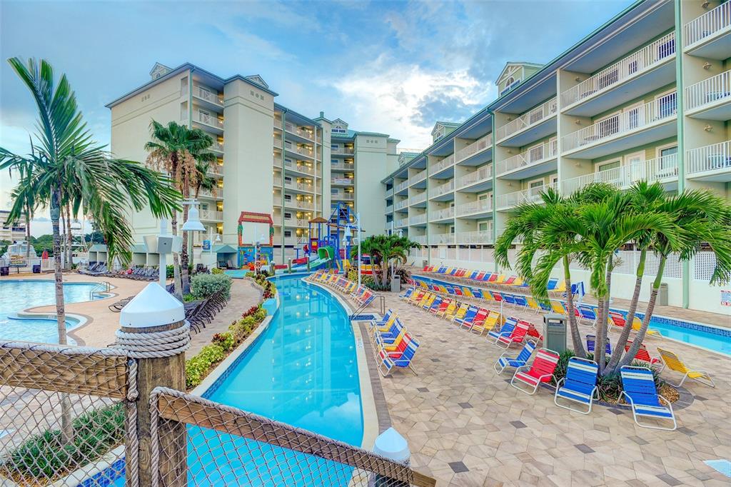 A resort pool area features a lazy river, lounge chairs, palm trees, and multi-story buildings — 399 2ND STREET UNIT 216, INDIAN ROCKS BEACH, FL