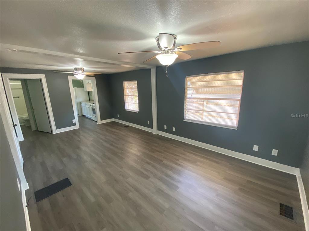 An empty room with gray walls, wood-look flooring, two ceiling fans, and a kitchen view — 227 E BARNES AVENUE, EUSTIS, FL