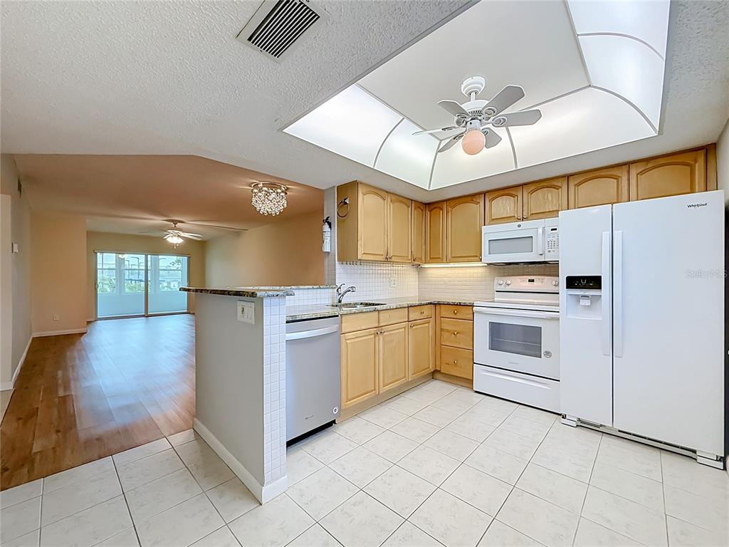 A kitchen with light wood cabinets, white appliances, and tile flooring opens to a living area — 952 VIRGINIA STREET UNIT 207, DUNEDIN, FL