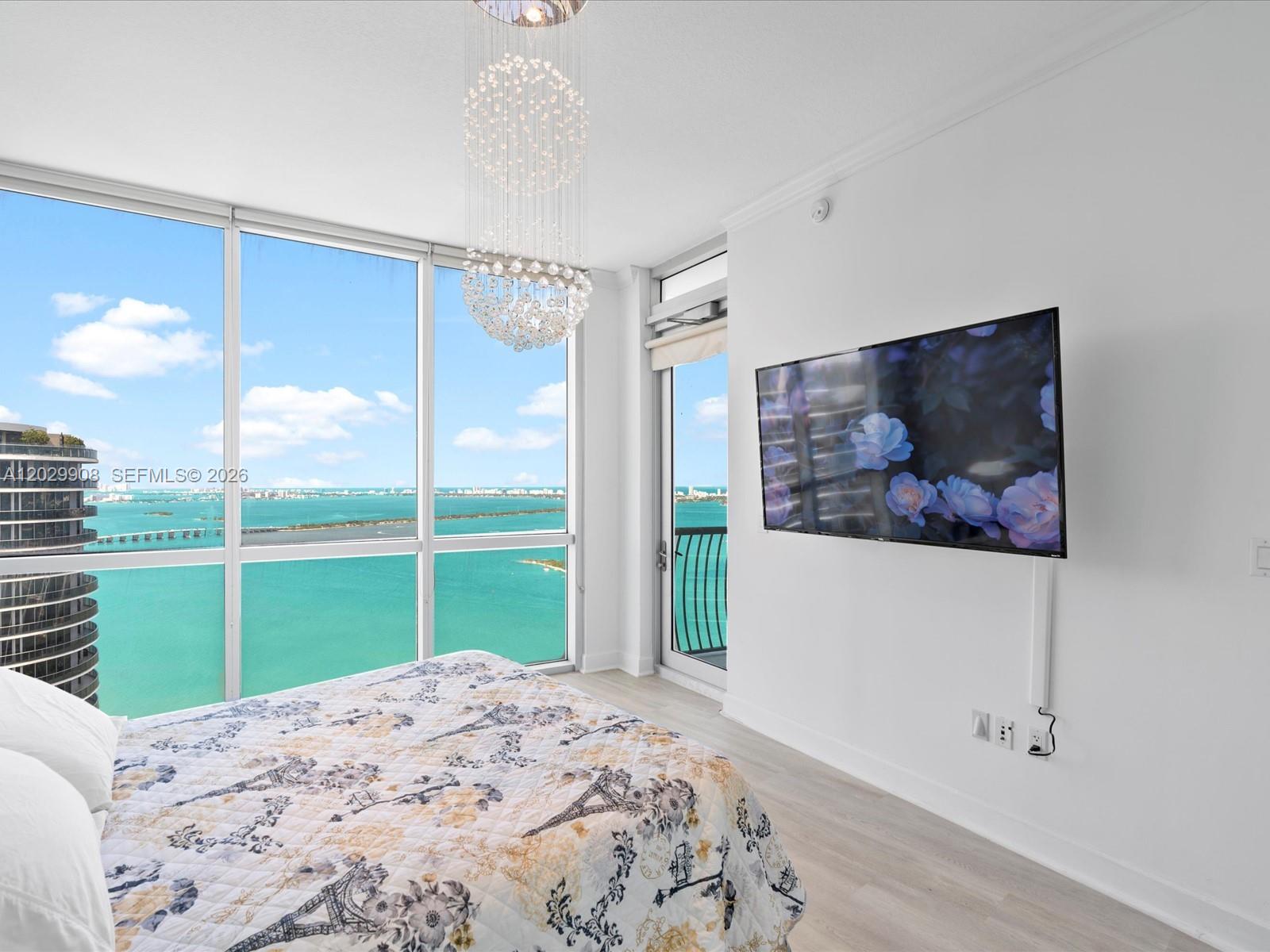 A bedroom features a crystal chandelier, a wall-mounted television, and floor-to-ceiling windows overlooking the ocean — 1750 N Bayshore Dr # 5601, Miami FL 33132, MIAMI, FL