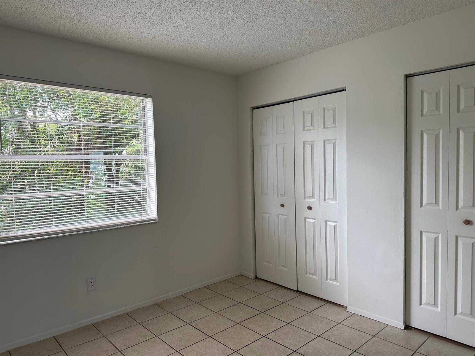 An empty room features white walls, beige tile flooring, a window with blinds, and closet doors — 4606 SE Binnacle Way # 1, Stuart FL 34997, STUART, FL