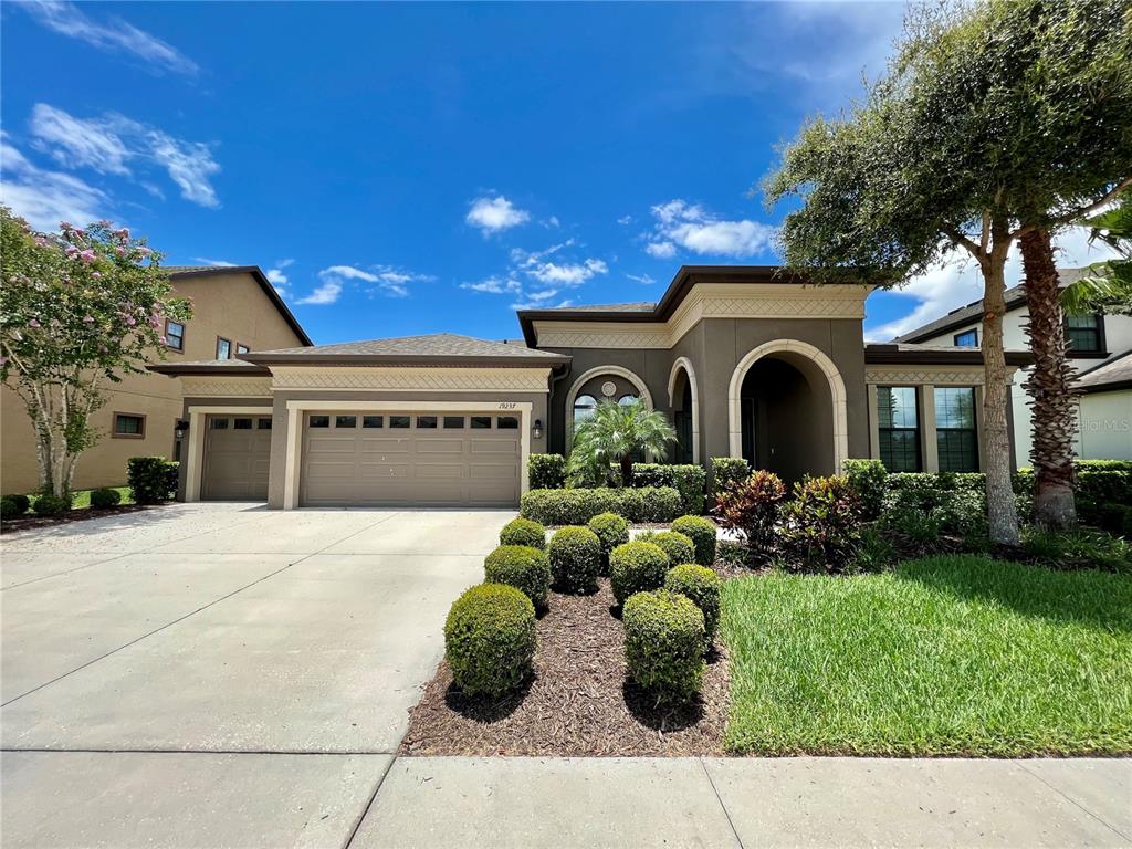 A single-story suburban house features a tan stucco exterior, a three-car garage, and manicured landscaping — 19237 FALCON CREST BOULEVARD, LAND O LAKES, FL