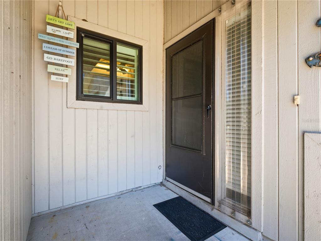 A concrete porch features a brown storm door, a window with blinds, and decorative wall signs — 4438 SEA MIST DRIVE UNIT 4438, NEW SMYRNA BEACH, FL