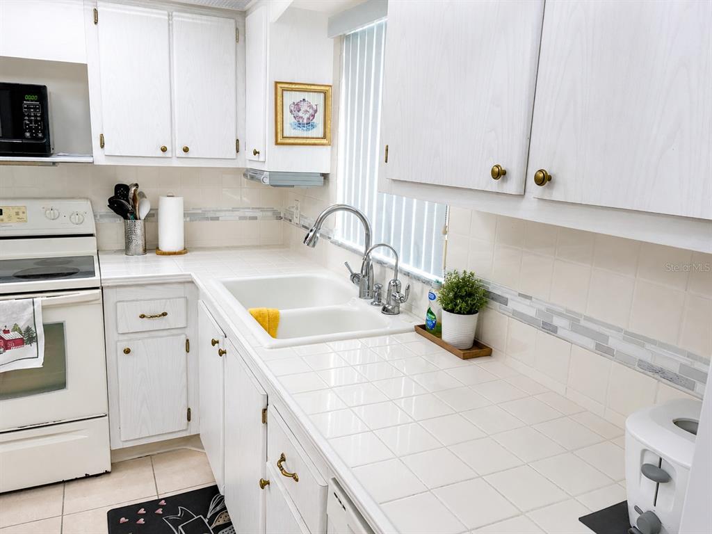 A kitchen features white cabinets, a tiled countertop, a double sink, and white appliances — 4575 COVE CIRCLE UNIT 506, ST PETERSBURG, FL